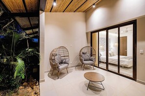 A modern interior with two wicker chairs, a round table, and a sliding glass door leading to a bedroom with a bed and a nightstand. - Charming Villa with Pool, Modern Amenities & Near Famous Surf Spots (Tamarindo)