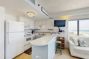 A modern kitchen with white cabinets, a central island, a built-in microwave, and a flat-screen TV mounted above the island. - Oceanfront studio with private balcony, pool & direct beach access (Myrtle Beach)
