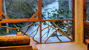 Una habitación con un amplio ventanal con vista a un río, muebles de madera y una lámpara colgante.