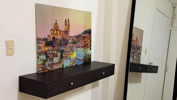 A room with a wooden floor, a black wall-mounted shelf with a colorful cityscape painting, a black trash bin, and a large mirror reflecting a door and a cabinet. - Encantador depa en Polanco, Ideal para viajeros. (Ciudad de México)