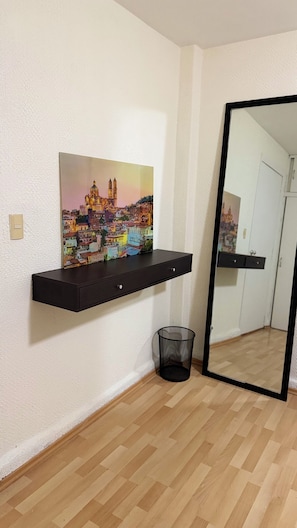 Ein Zimmer mit Holzboden, einer an der Wand befestigten schwarzen Ablage mit einem farbenfrohen Stadtbild und einer schwarzen Mülltonne. Ein großes Spiegelbild zeigt eine Tür und einen Schrank.