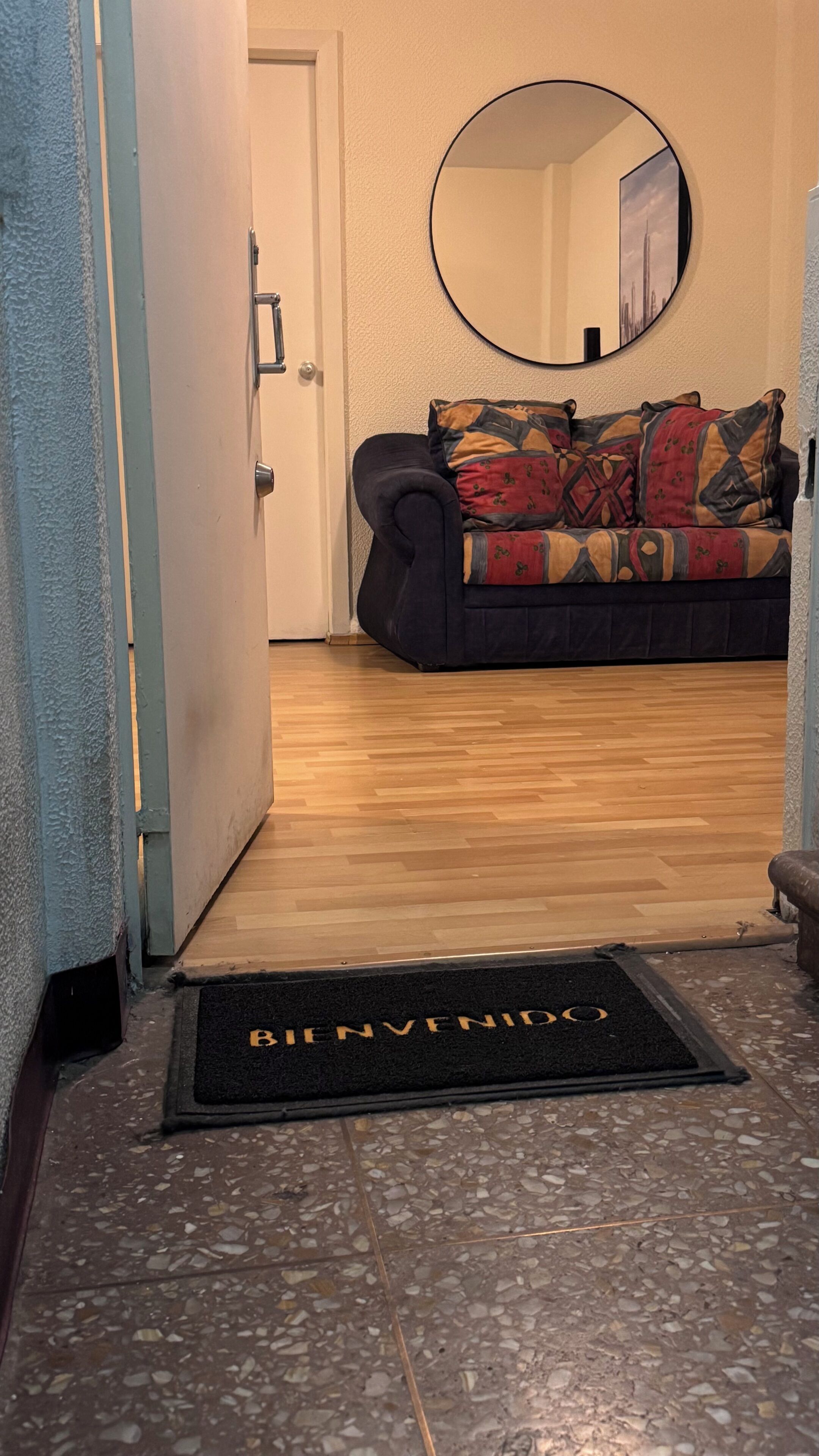 A hallway leading to a room with a patterned sofa, a round mirror on the wall, and a welcome mat with 'BEN VENIDO' text.