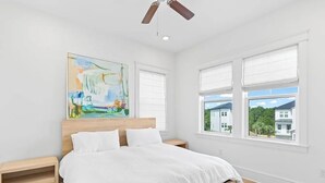 Une chambre à coucher comprenant un lit, une tête de lit en bois, deux tables de chevet, un ventilateur de plafond et une grande fenêtre avec des stores.