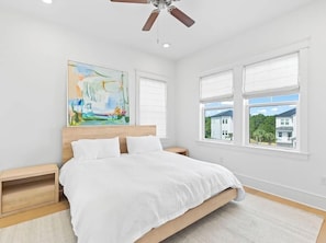 Ein Schlafzimmer mit einem Bett, einem Holz-Kopfteil, zwei Nachttischen, einem Deckenventilator und einem großen Fenster mit Jalousien.