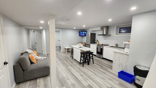 A modern kitchen with a central island, a dining area with a sofa and chairs, and a flat-screen TV mounted above the island. - Newly renovated 2 bedroom basement with seperate entrance. (Whitby)