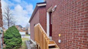 A residential building with a brick exterior, a wooden staircase leading to a door, and a small garden area with a tree and stepping stones. - Newly renovated 2 bedroom basement with seperate entrance. (Whitby)