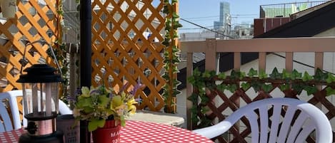 Een tafel met een rood-wit geruit tafelkleed, een Coleman campinglantaarn en een bloempot op een balkon met een houten lattenhek en witte stoelen.