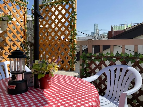 A table with a red and white checkered tablecloth, a Coleman camping lantern, and a potted plant on a balcony with a wooden lattice fence and white chairs. - The Familiar Inn-Hotel and Inn Business (Osaka)