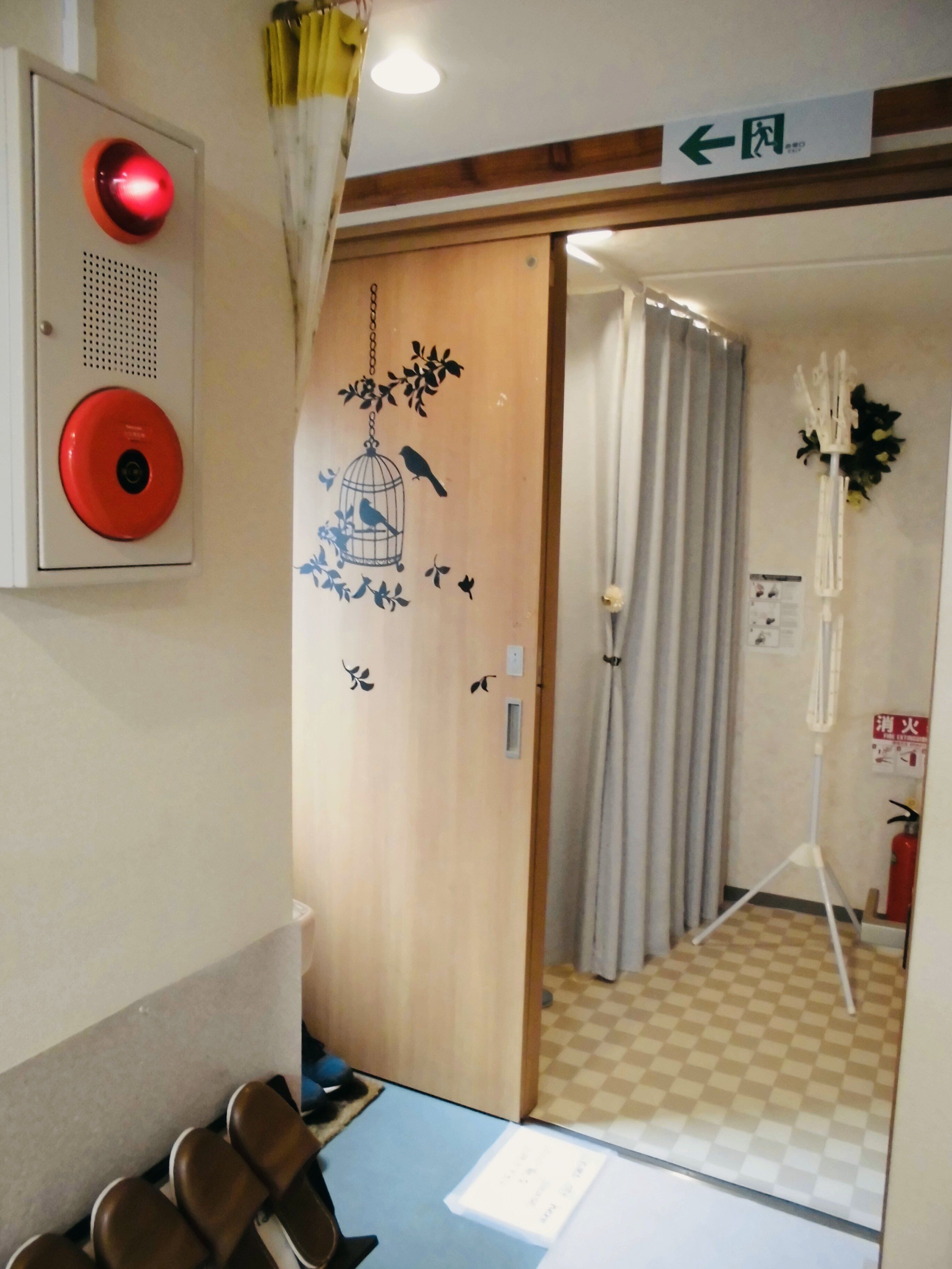 A hotel room with a wooden door, a red emergency alarm, a blue table with a chair, and a checkered floor.