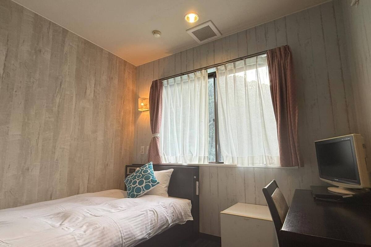 A bedroom with a bed, a desk with a computer, a window with curtains, and a wall-mounted light.
