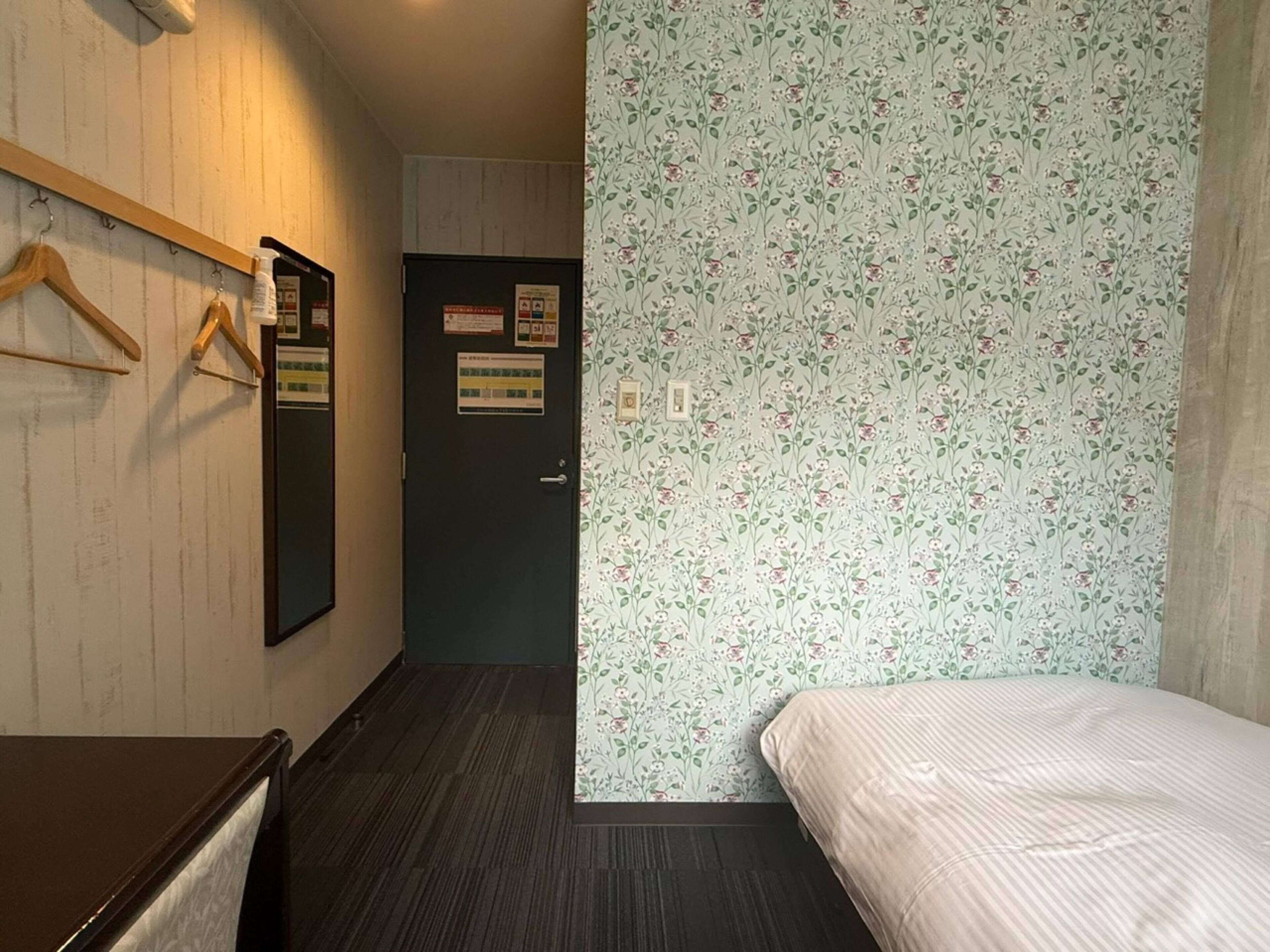 A hotel room with a bed, a desk, a mirror, a wooden hanger rack, and a door with a floral wallpaper.