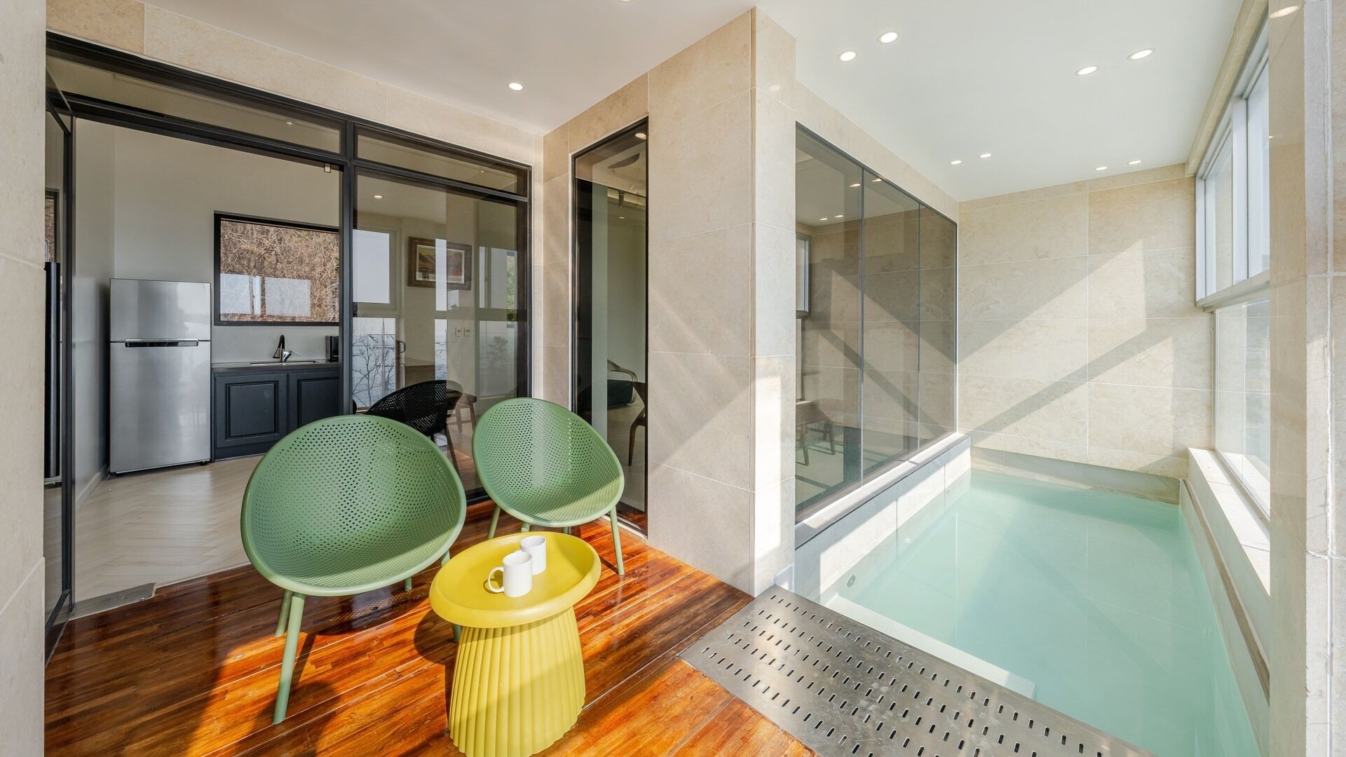 A modern interior with a wooden floor, two green chairs, a yellow stool, a glass-enclosed pool, and a kitchen area visible through glass doors.