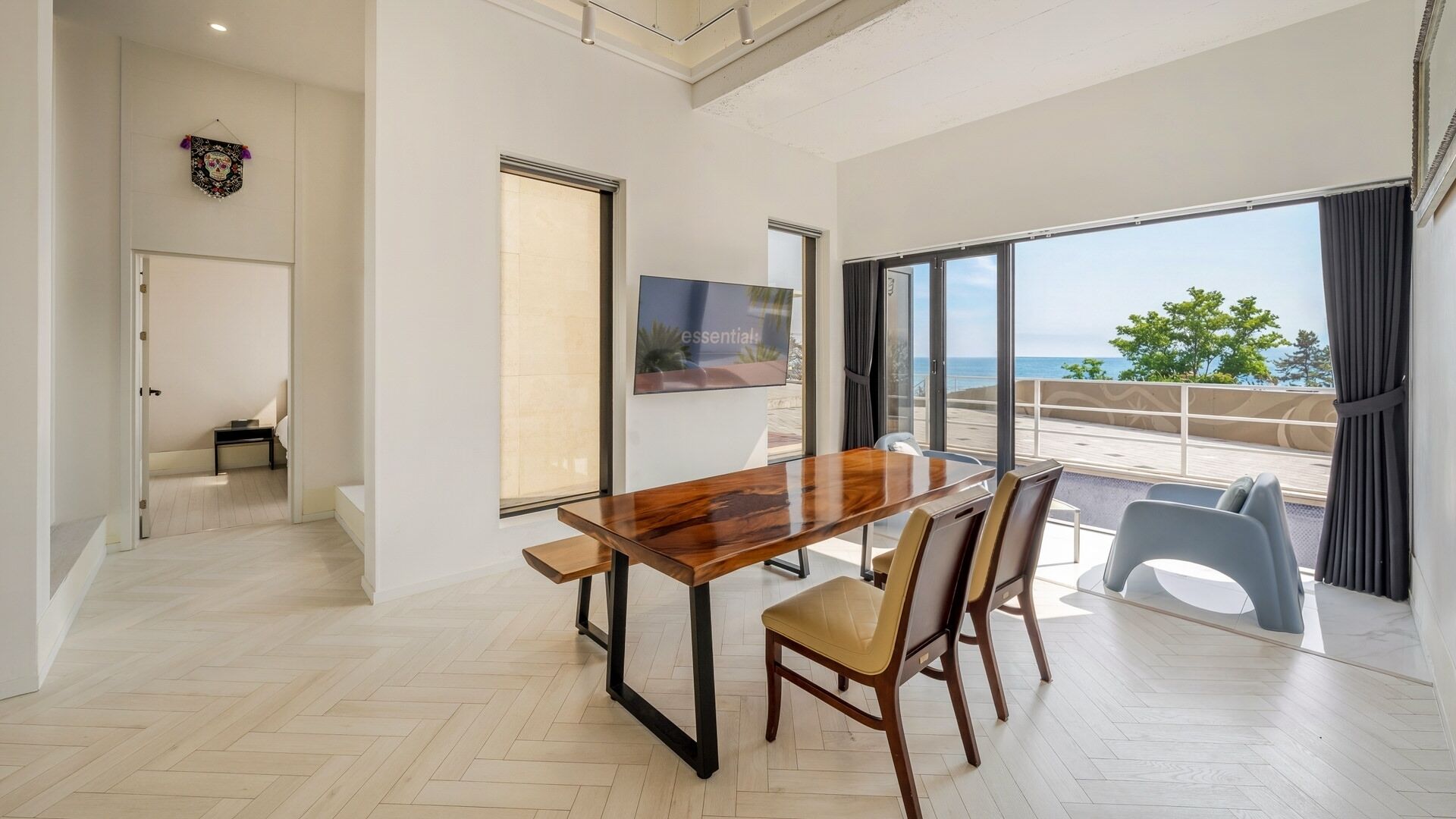 A modern dining area with a wooden table and chairs, a television, and a view of the sea through large windows.