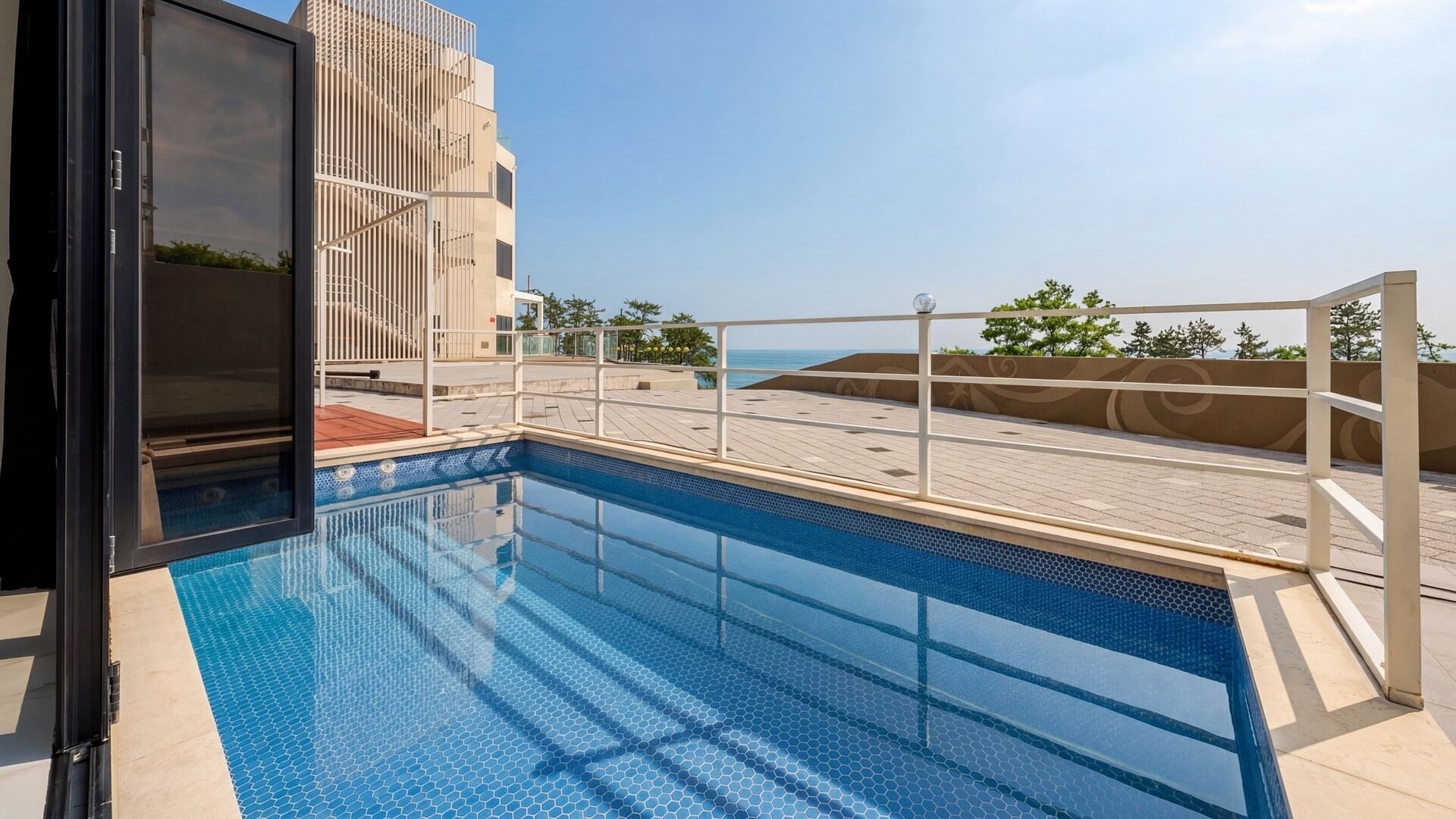 A rooftop swimming pool with a clear view of the sea, surrounded by a modern building with a glass facade.