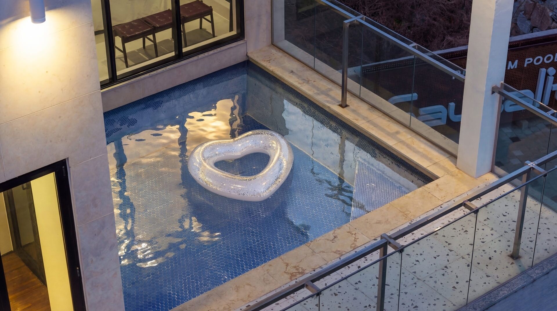 An indoor swimming pool with a heart-shaped float, glass railings, and a view of a building with benches outside.