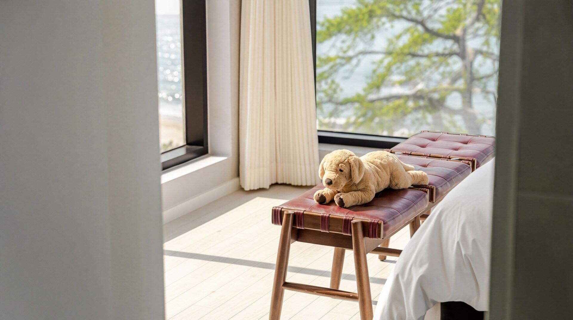 Une pièce avec une grande fenêtre, un jouet en peluche posé sur un banc en cuir et un lit avec une literie blanche.