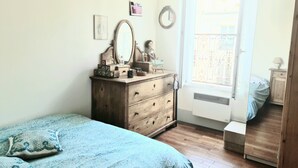 Ein Schlafzimmer mit einem Holzkontoi, einem Bett mit gemusterten Bettbezügen, einem Fenster mit Aussicht und einem kleinen Nachttisch mit Lampe.