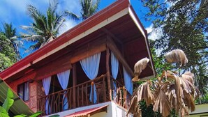 Une maison à deux étages dotée d’un balcon, entourée d’une végétation luxuriante et de plantes tropicales.