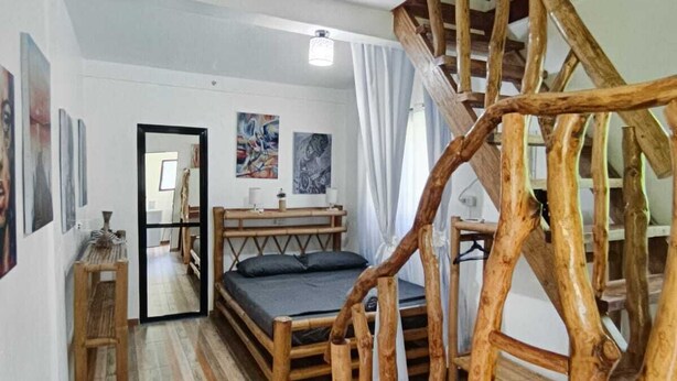 Une pièce avec un escalier en bois, un lit doté d’un matelas sombre et un miroir qui reflète la pièce.