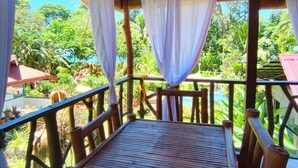 Une terrasse en bois, agrémentée d’une table et de chaises, entourée d’une balustrade en bambou, et offrant une vue sur un jardin luxuriant et une piscine.