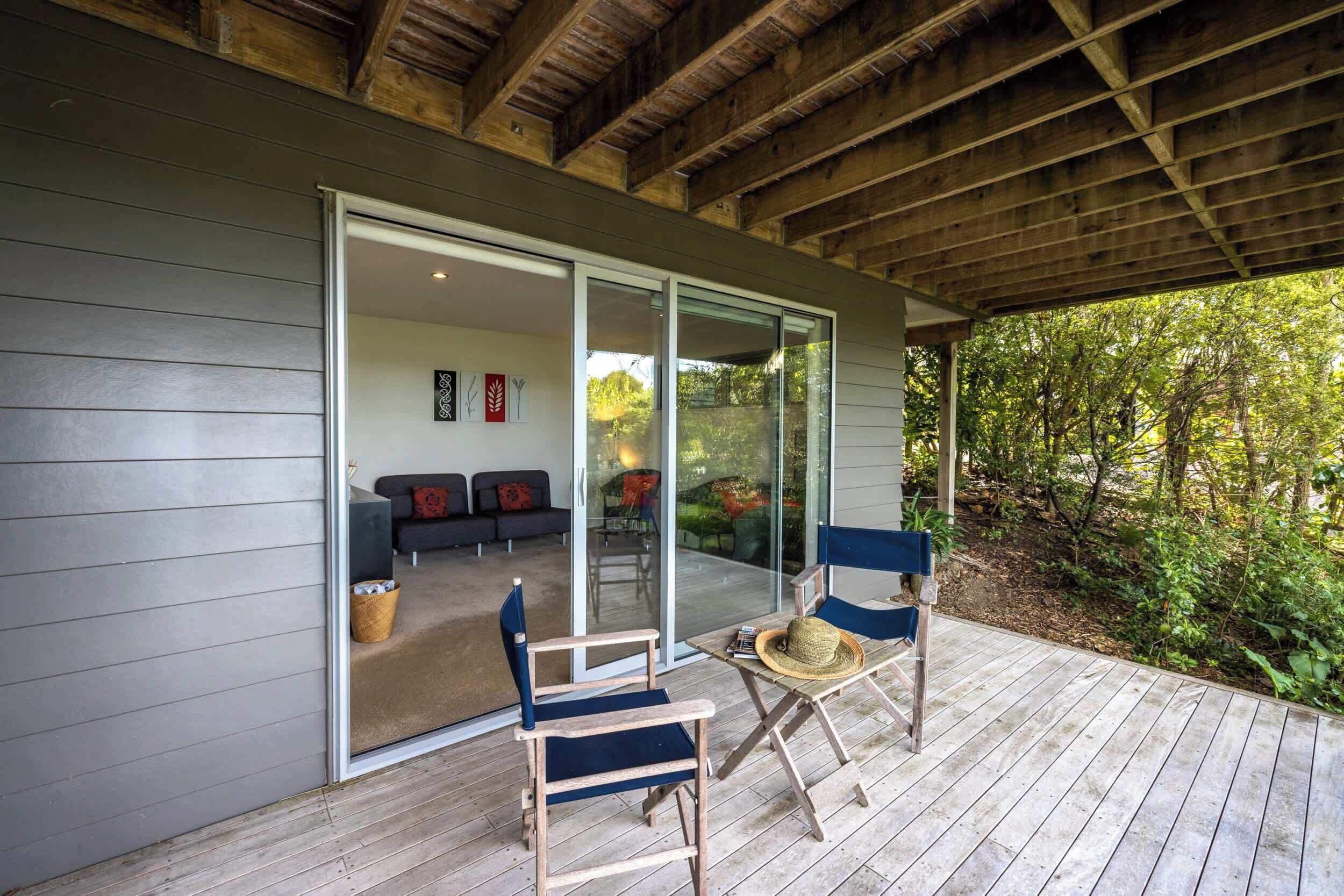 A covered patio with wooden flooring, two wooden chairs, a woven basket, and a glass door leading to an interior space with a sofa and cushions.
