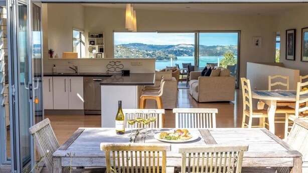 A modern kitchen with a dining area, a view of the sea, and a large glass door leading to the outside. - Wakainga — Church Bay Estate (Waiheke Island)