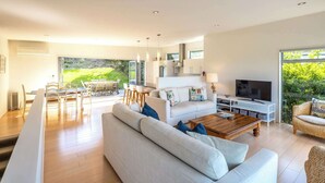 A modern living room with a sofa, coffee table, TV, and dining area. - Wakainga — Church Bay Estate (Waiheke Island)
