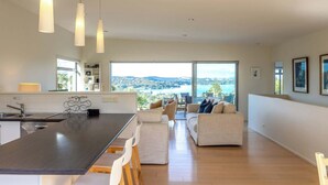 A modern kitchen with a central island, a dining area with white chairs, and a sofa set. - Wakainga — Church Bay Estate (Waiheke Island)