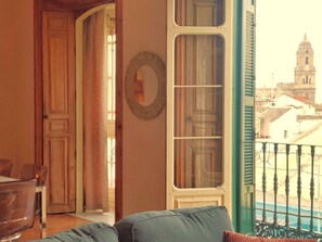 Ein Zimmer mit Blick auf ein Gebäude durch ein Fenster, eine Tür, die in einen anderen Raum führt, und ein wandmontierter Spiegel.