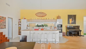 A modern kitchen with a central island, bar stools, a piano, and a dining area with a table and chairs. - Villa La Carqueiranaise (CARQUEIRANNE)