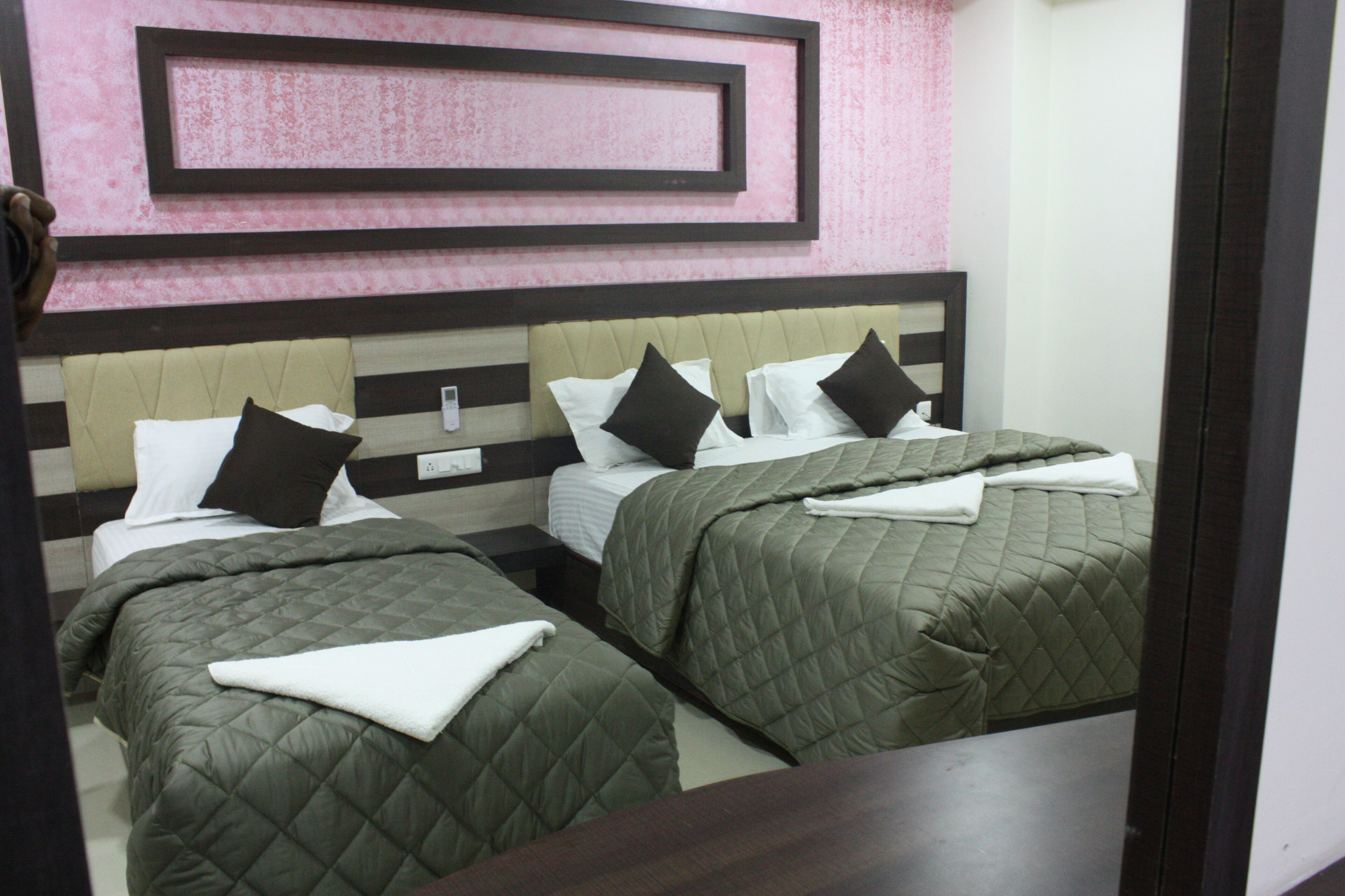 Une chambre d’hôtel avec deux lits, chacun recouvert d’une courtepointe verte matelassée, avec du linge de lit blanc et des oreillers foncés. La chambre présente un design moderne, avec un panneau mural rose et marron et une tête de lit en bois.
