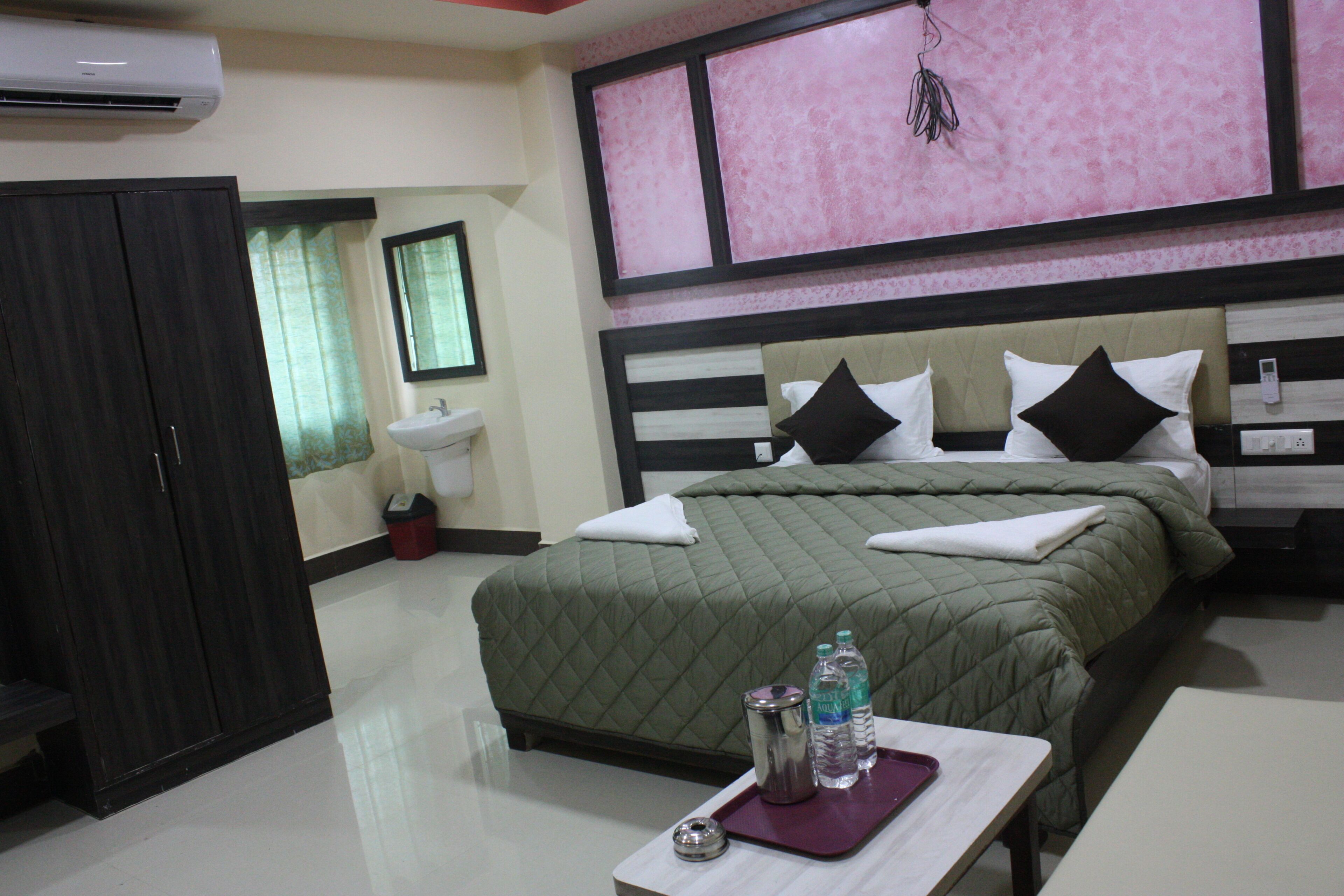 A hotel room with a bed, a bedside table with water bottles, a wardrobe, a sink, and a mirror.