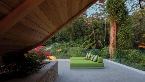 Un área de estar moderna al aire libre, con un sofá y cojines verdes, rodeada de abundante vegetación y un muro de piedra.