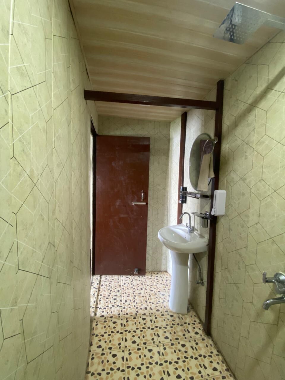 A narrow bathroom with a tiled floor, a white sink, a mirror, and a wooden door.