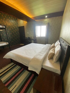 A hotel room with a large bed, a wooden headboard, a striped rug, a window with curtains, and a small table with chairs. - Stone Rose Vista (Tehri)