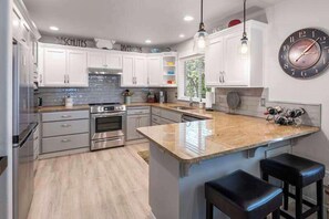 A modern kitchen with white cabinets, stainless steel appliances, a large island with bar stools, and a wall clock. - Deschutes Riverhaus | Luxury Retreat, Hot Tub (Sunriver)