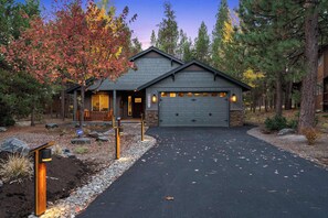 A two-story house with a garage, surrounded by trees and a gravel driveway. - Deschutes Riverhaus | Luxury Retreat, Hot Tub (Sunriver)
