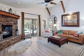 A living room with a stone fireplace, a ceiling fan, a sofa, and a painting on the wall. - Deschutes Riverhaus | Luxury Retreat, Hot Tub (Sunriver)