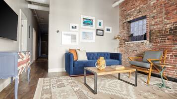A living room with a blue sofa, a coffee table, and a brick wall. There are framed pictures on the wall and a television mounted on the wall.