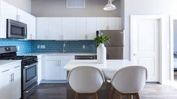 Cozinha moderna com armários brancos, geladeira de aço inoxidável, micro-ondas e fogão. Apresenta um painel de azulejos azuis e uma ilha branca com duas cadeiras.