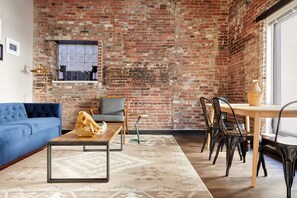 A room with a brick wall, a blue sofa, a wooden coffee table, a grey armchair, and a dining table with chairs. - Kasa At Boston Common (Boston)