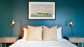 A bedroom with a large bed, two bedside tables, a framed painting of a sailing ship, and wall-mounted lamps.