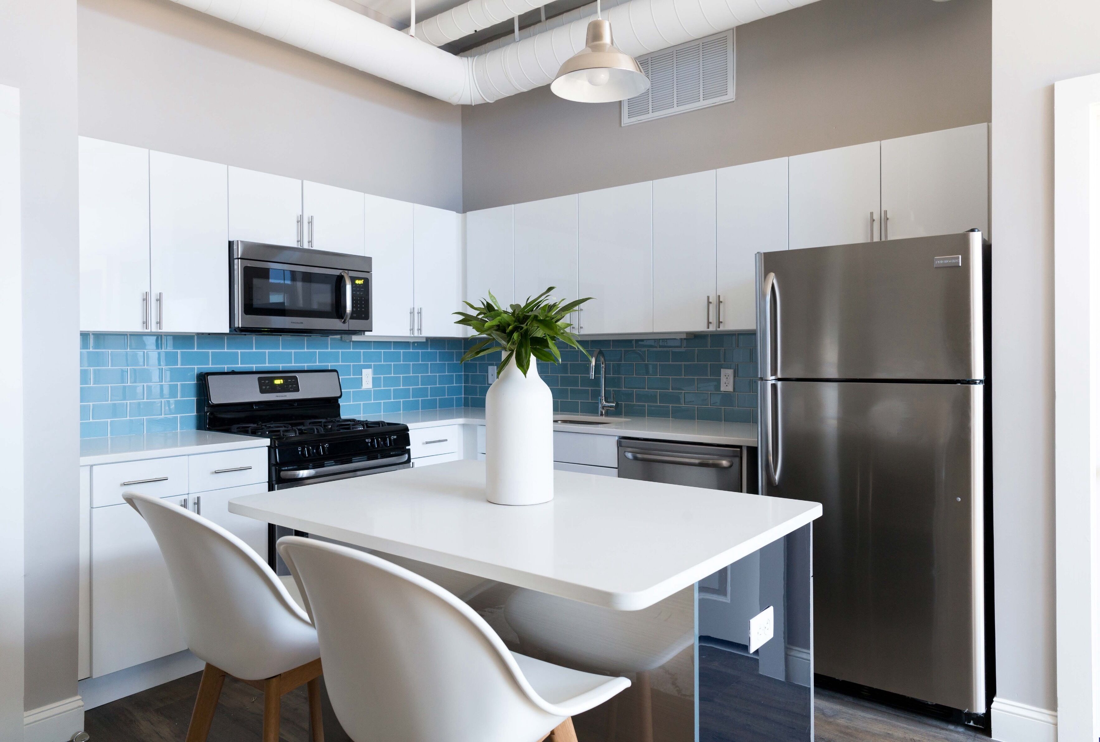 A modern kitchen with white cabinets, a stainless steel refrigerator, a microwave, and a stove. There is a white island with two chairs and a potted plant.