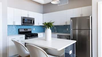 A modern kitchen with white cabinets, a stainless steel refrigerator, a microwave, and a stove. There is a white island with two chairs and a potted plant.