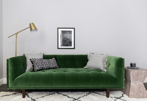 Un sofá verde con dos almohadas, un cuadro enmarcado en la pared, una lámpara de pie y una pequeña mesita de madera con un vaso.
