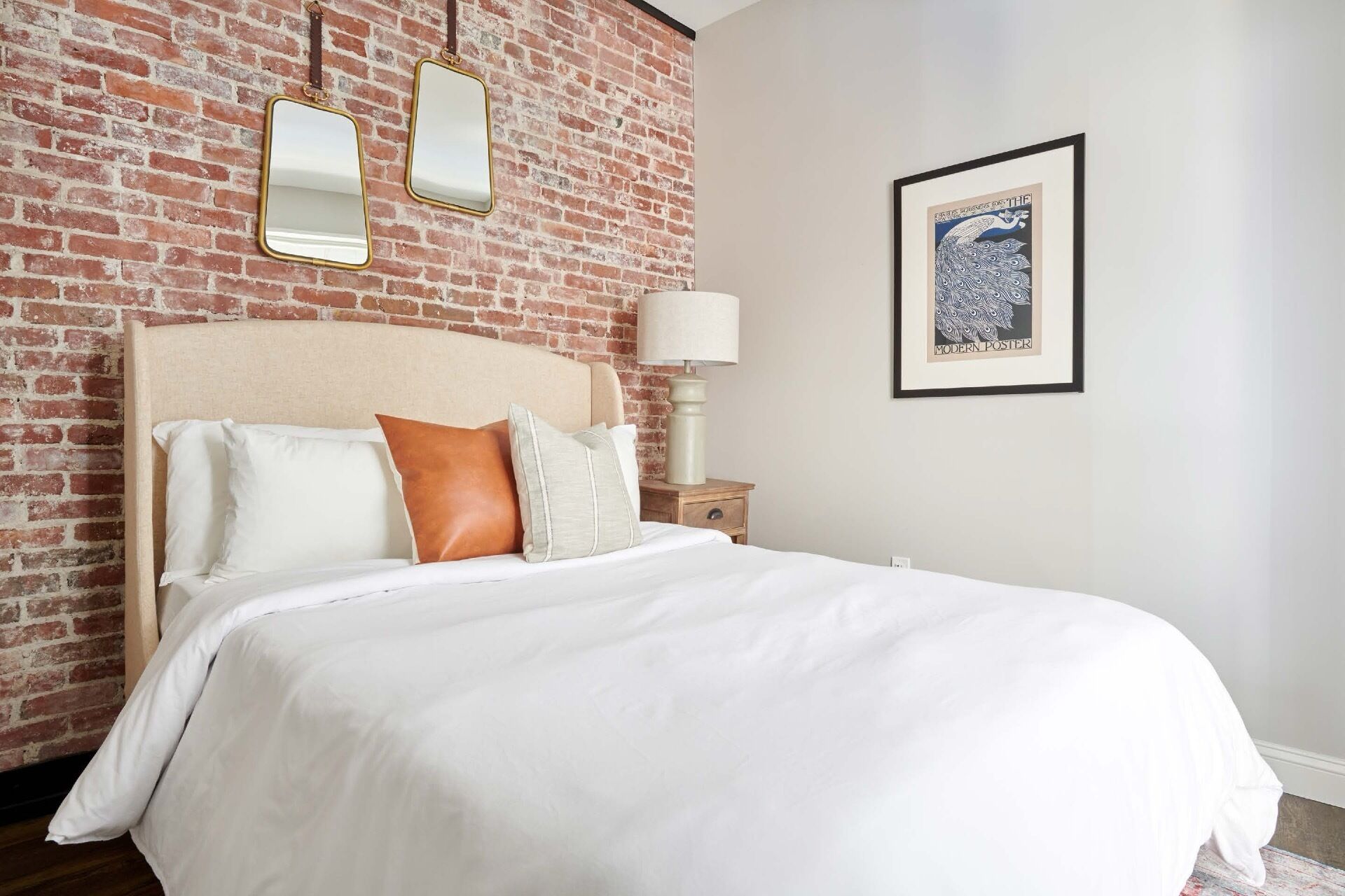 Une chambre d’hôtel avec un mur en briques, un lit avec une literie blanche et un oreiller marron, une table de chevet avec une lampe, et un tableau encadré accroché au mur.