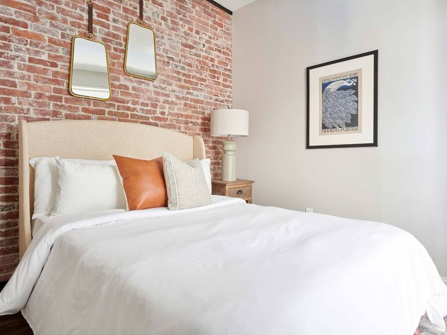 Une chambre d’hôtel avec un mur en briques, un lit avec une literie blanche et un oreiller marron, une table de chevet avec une lampe, et un tableau encadré accroché au mur.
