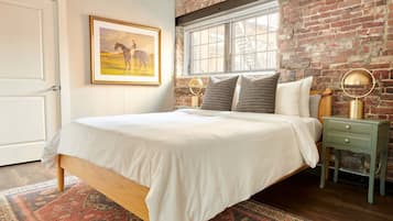 A bedroom with a bed, a window, a brick wall, a framed picture, and a nightstand with a lamp.