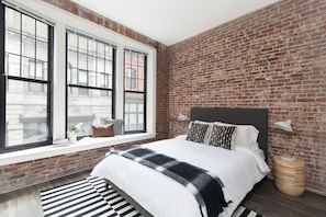A bedroom with a brick wall, a large window, a bed with white bedding, and a bedside lamp. - Kasa At Boston Common (Boston)