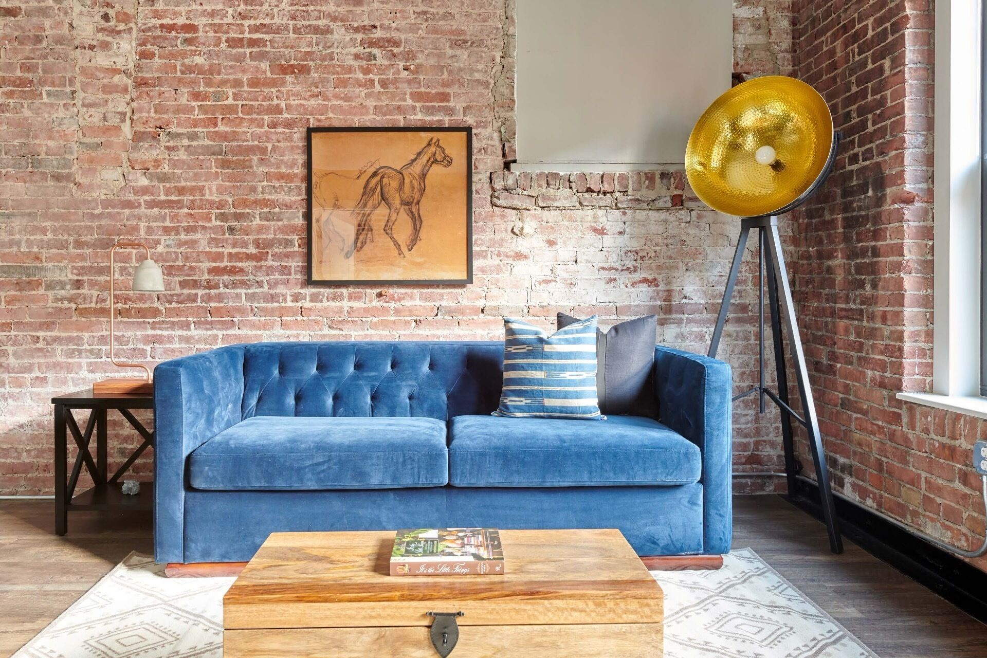 Une pièce avec un canapé bleu, une table basse en bois, un tableau encadré représentant un cheval, un lampadaire et un mur en briques.